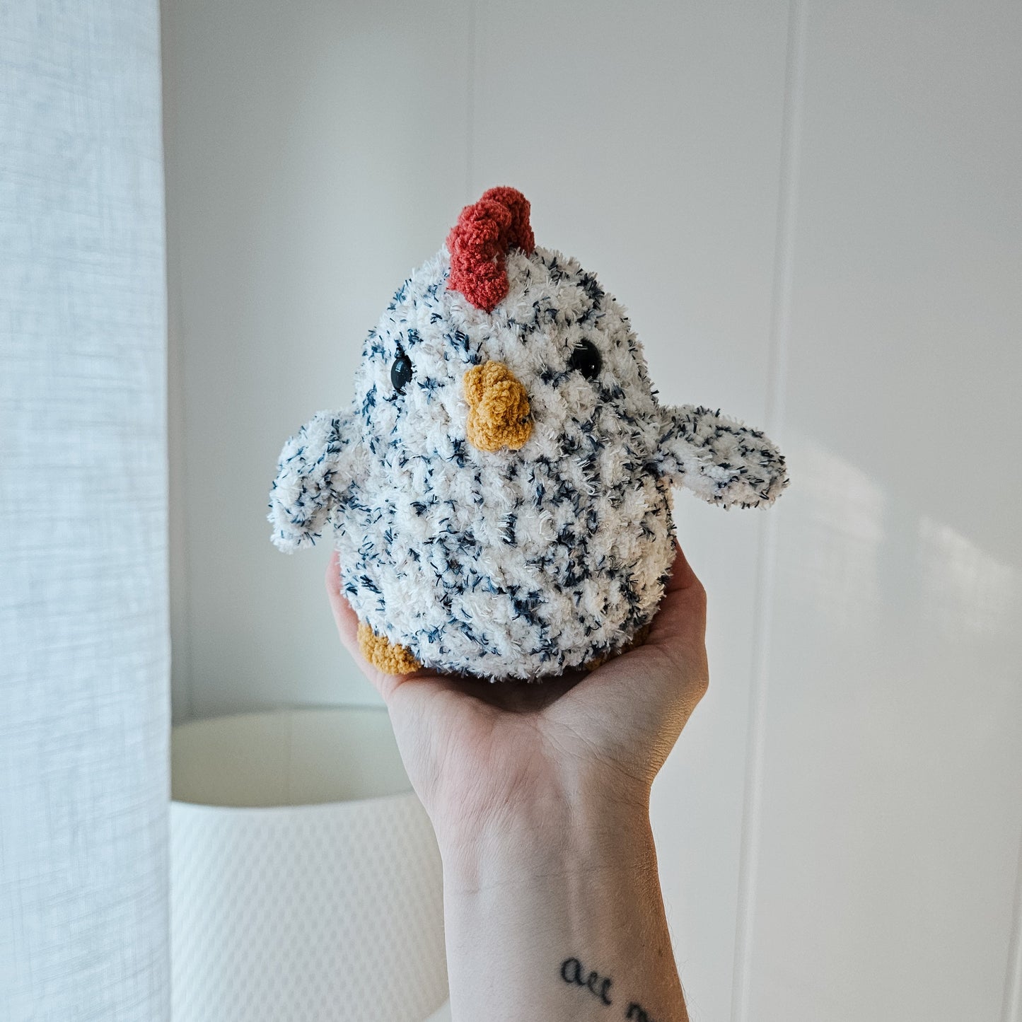 A crochet pattern for a chicken-shaped Chubby Buddy held in a person's hand, showing the chicken with a gray body, white and orange details, and a red comb.