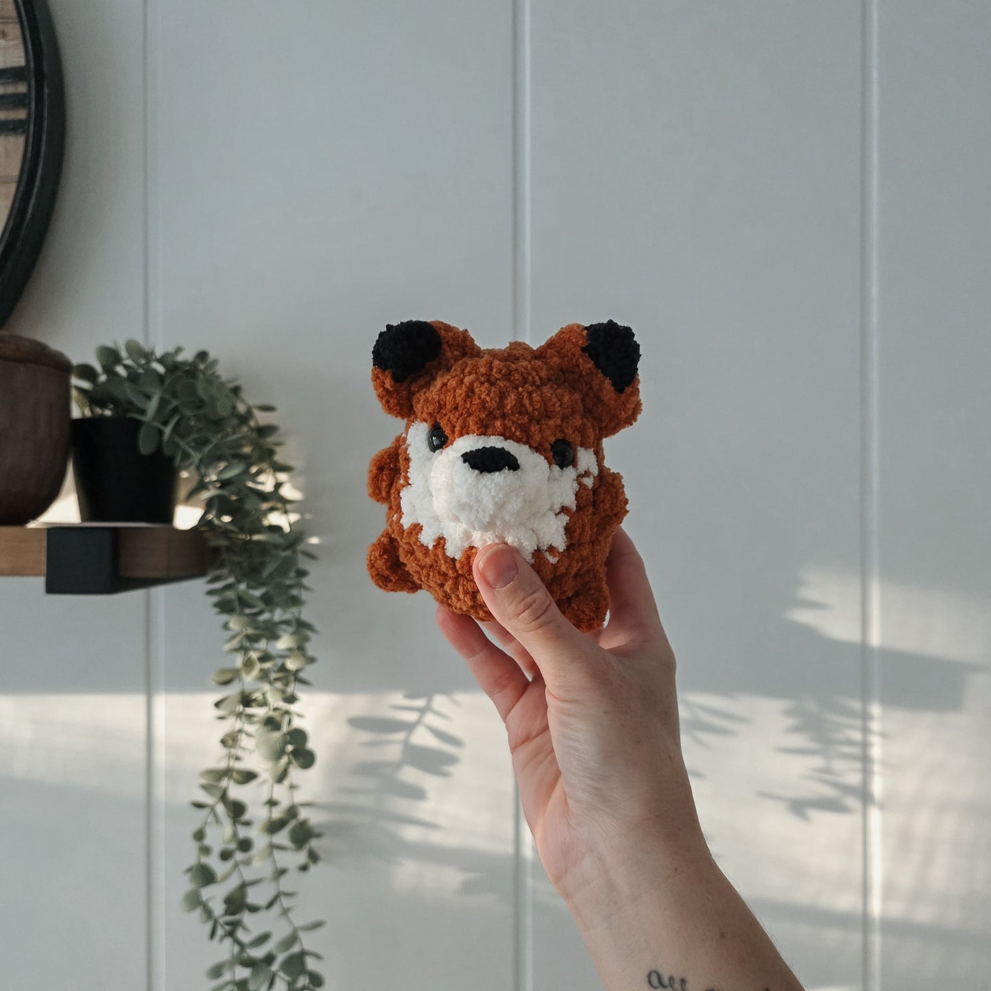 Hand holding a small crochet orange fox toy against a light wooden panel background