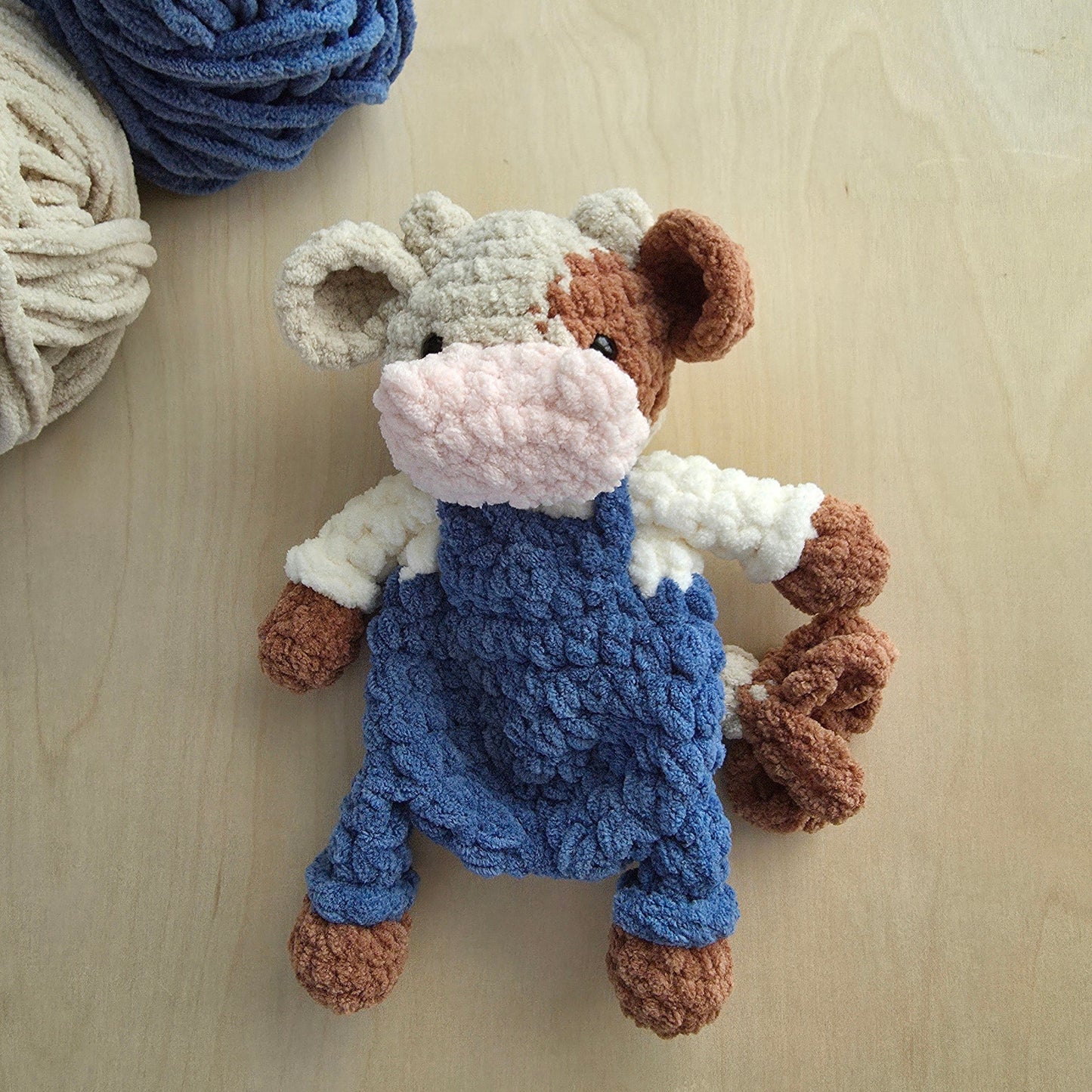 A crocheted cow plush toy wearing blue overalls and a white shirt, placed on a light wood surface with balls of yarn in the top left corner.