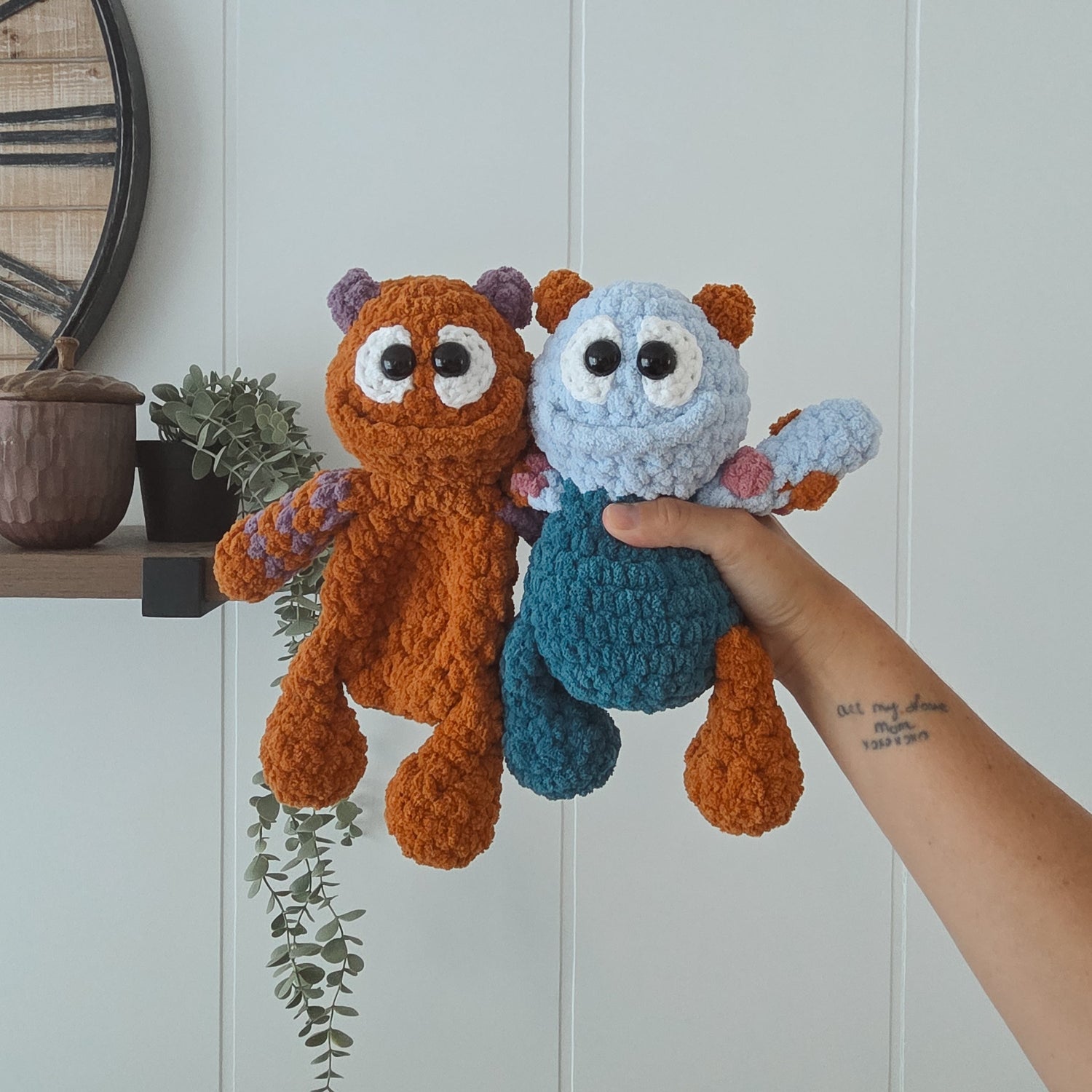 Two handmade crocheted plush toys are held up by a person's hand against a white paneled wall. The toy on the left is orange with purple ears and a striped scarf, and the toy on the right is light blue with a teal body and pink scarf.