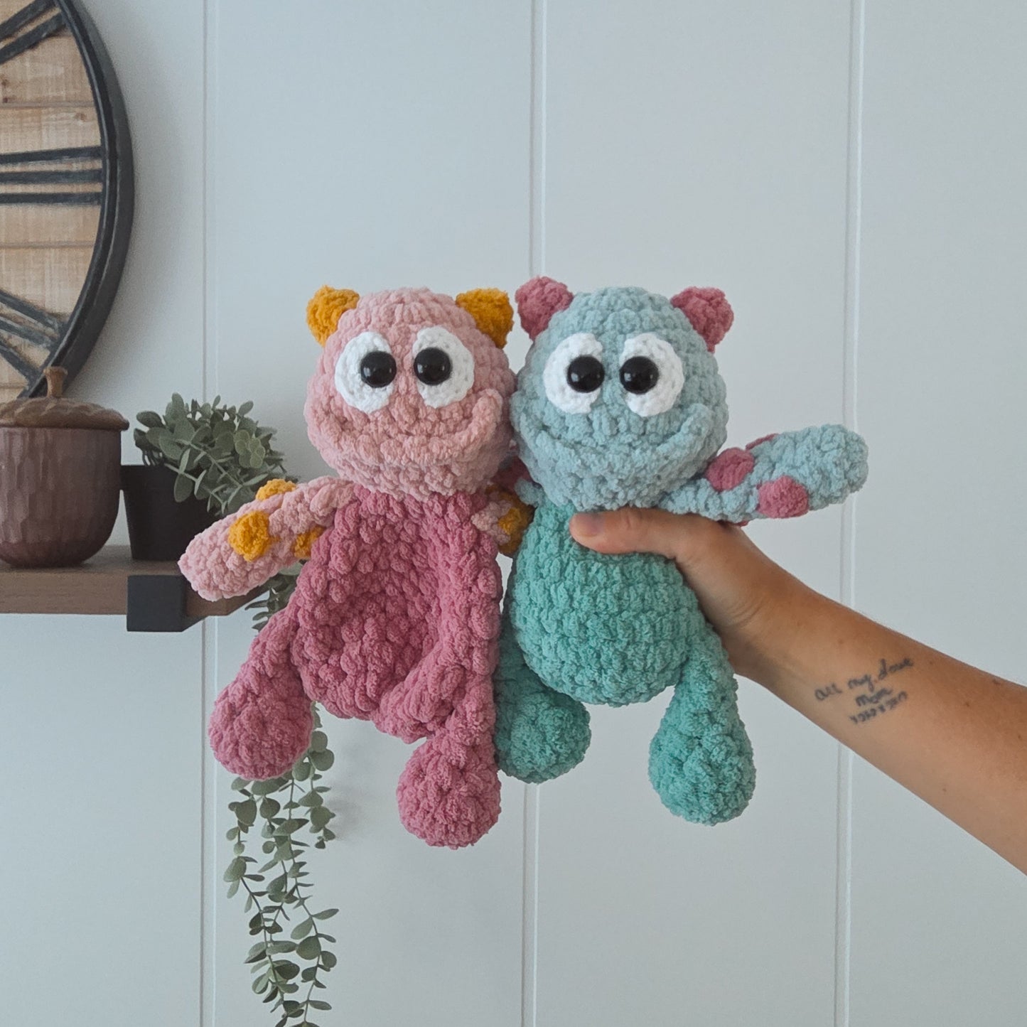 Two crocheted plush monster toys are held up by a person's hand against a light paneled wall. One toy is pink with yellow ears and the other is teal with pink ears.