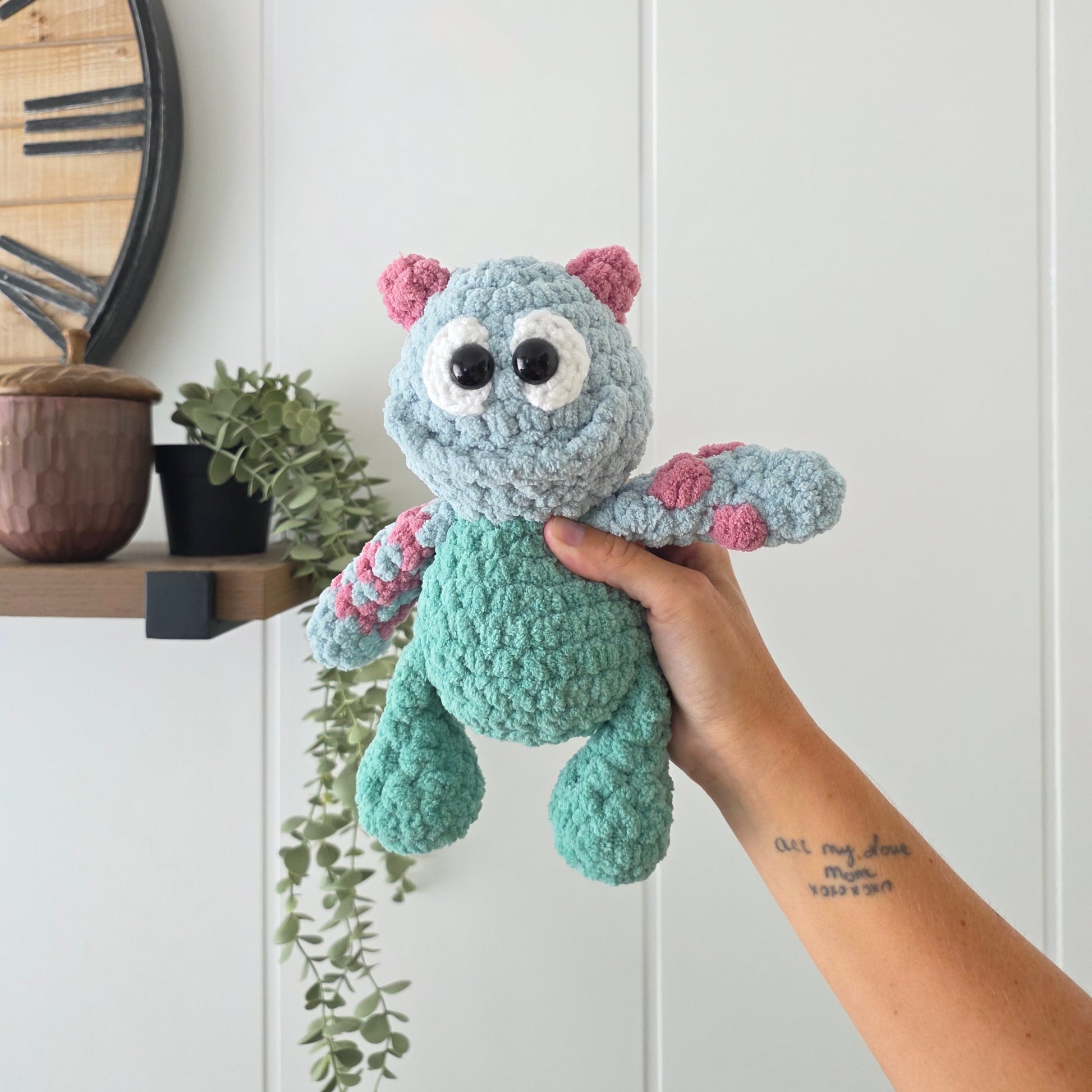 A handmade crocheted monster plush toy being held by a person's hand in front of a white paneled wall. The plush is light blue with a teal-green belly, pink ears, and pink-and-white striped arms.