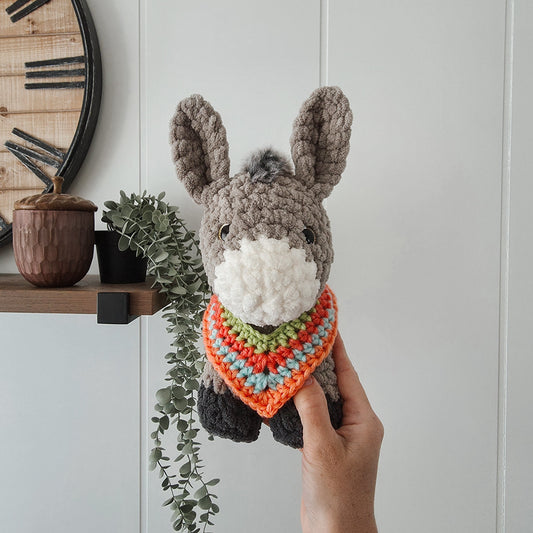 Hand holding a crochet donkey toy with a colorful scarf in a room with a clock and plants.