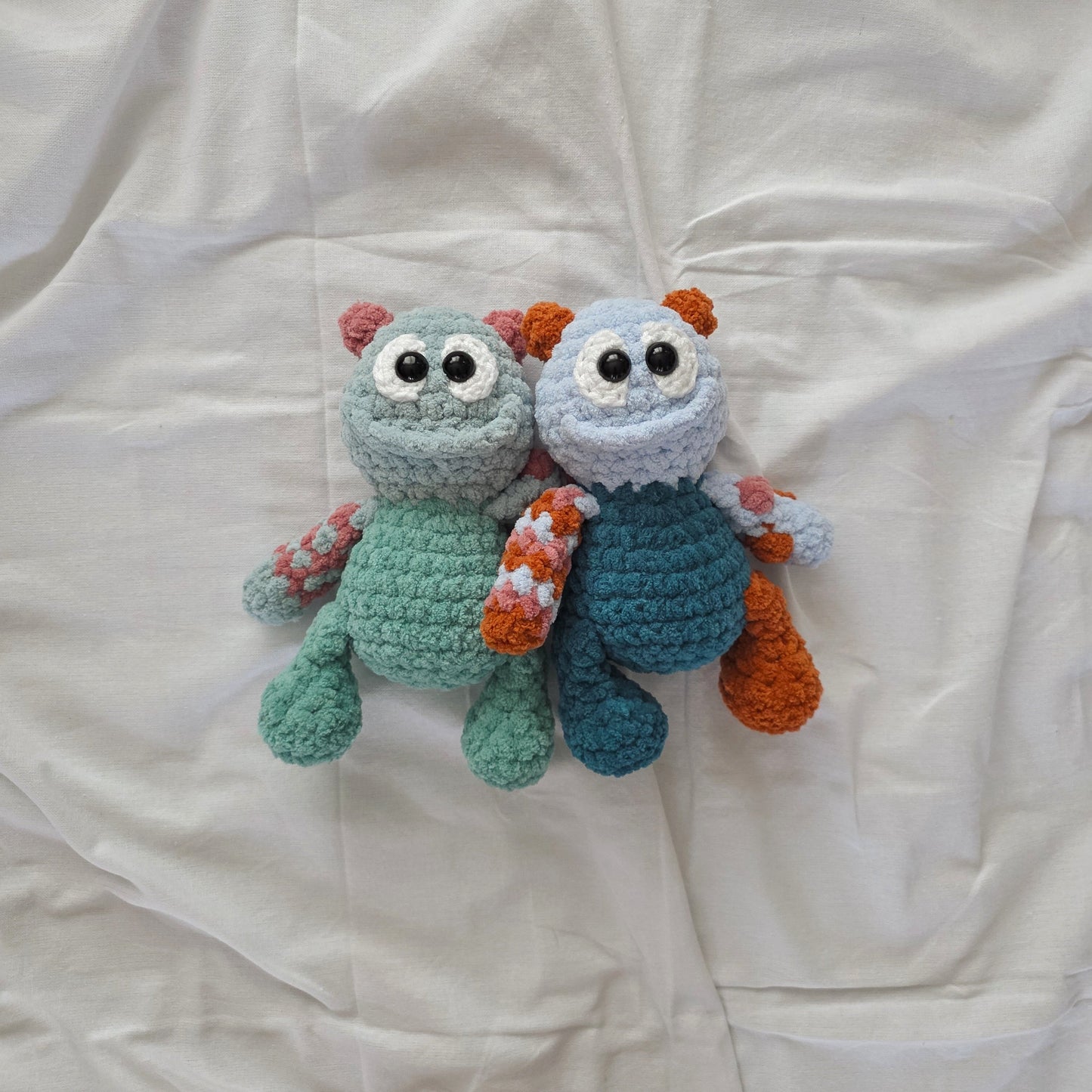 Two handmade crocheted monster toys lying on a wrinkled white fabric background. One toy is light blue-green with orange accents, and the other is dark blue with orange accents.